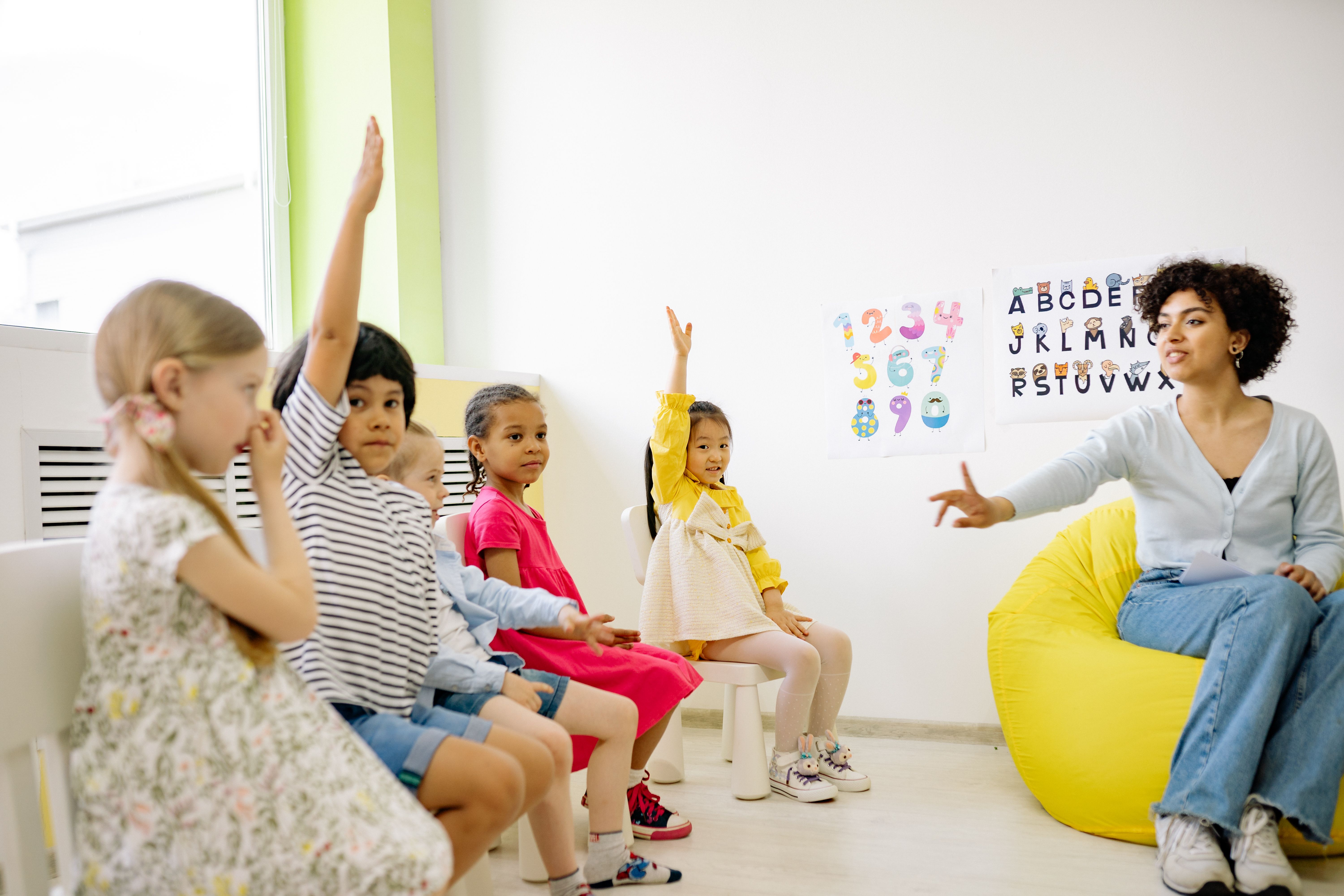 Teacher sitting with preschool children in a bright classroom, children raising hands eagerly